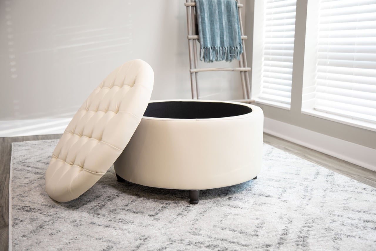 A Wovenbyrd Large Round Pintucked Storage Ottoman with Lift Off Lid (SKU: 7HSO0001 - FL0004) in cream faux leather featured in the corner of a living room.