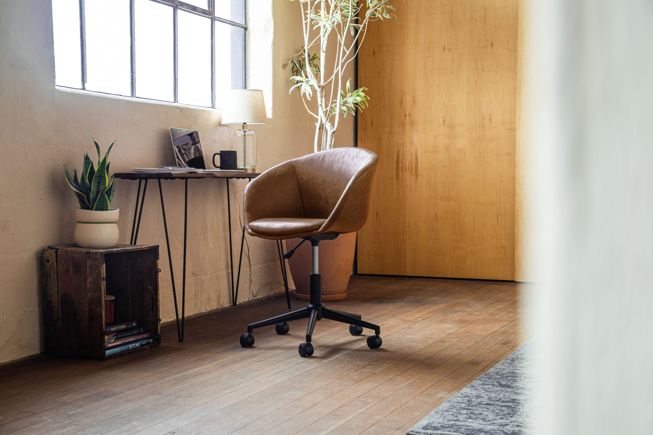 A Wovenbyrd Modern Curved Back Barrel Office Chair in Light Brown Faux Leather sits in a sunny, modern work-from-home office space
