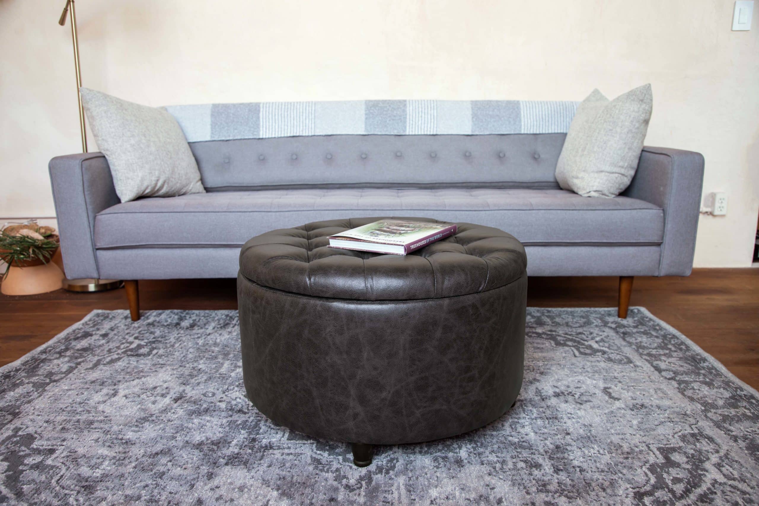 A Wovenbyrd Large Round Pintucked Storage Ottoman with Lift Off Lid sits in front of a couch in a living room, providing a foot rest and additional storage space.